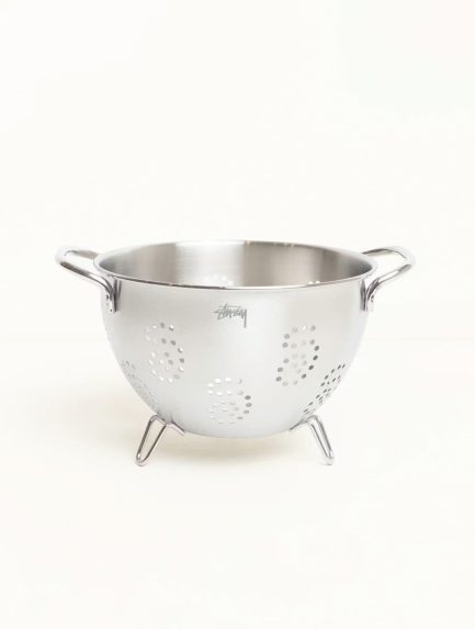 A stainless steel Stüssy strainer with an etched logo next to a branded glass pour over coffee server.