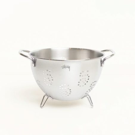 A stainless steel Stüssy strainer with an etched logo next to a branded glass pour over coffee server.