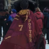 Dominic McLaughlin seen from behind wearing a red and gold Gryffindor Quidditch cloak carrying a broomstick and walking toward an outdoor stadium with Gryffindor and Hufflepuff flags.